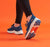 A person in black athletic leggings and white sneakers jumps against a bright orange background, with a large ARCO logo graphic at the bottom left.